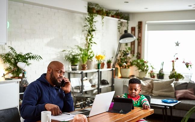 Adult man working from home, using phone at dining room table with laptop, with a small young boy sitting patiently with device
