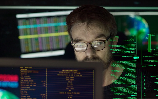 Close-up photo of a mature man surrounded by monitors & a holographic display which he is reading