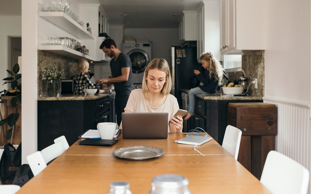 Adult woman holding mobile phone while using laptop at table with family in kitchen at home