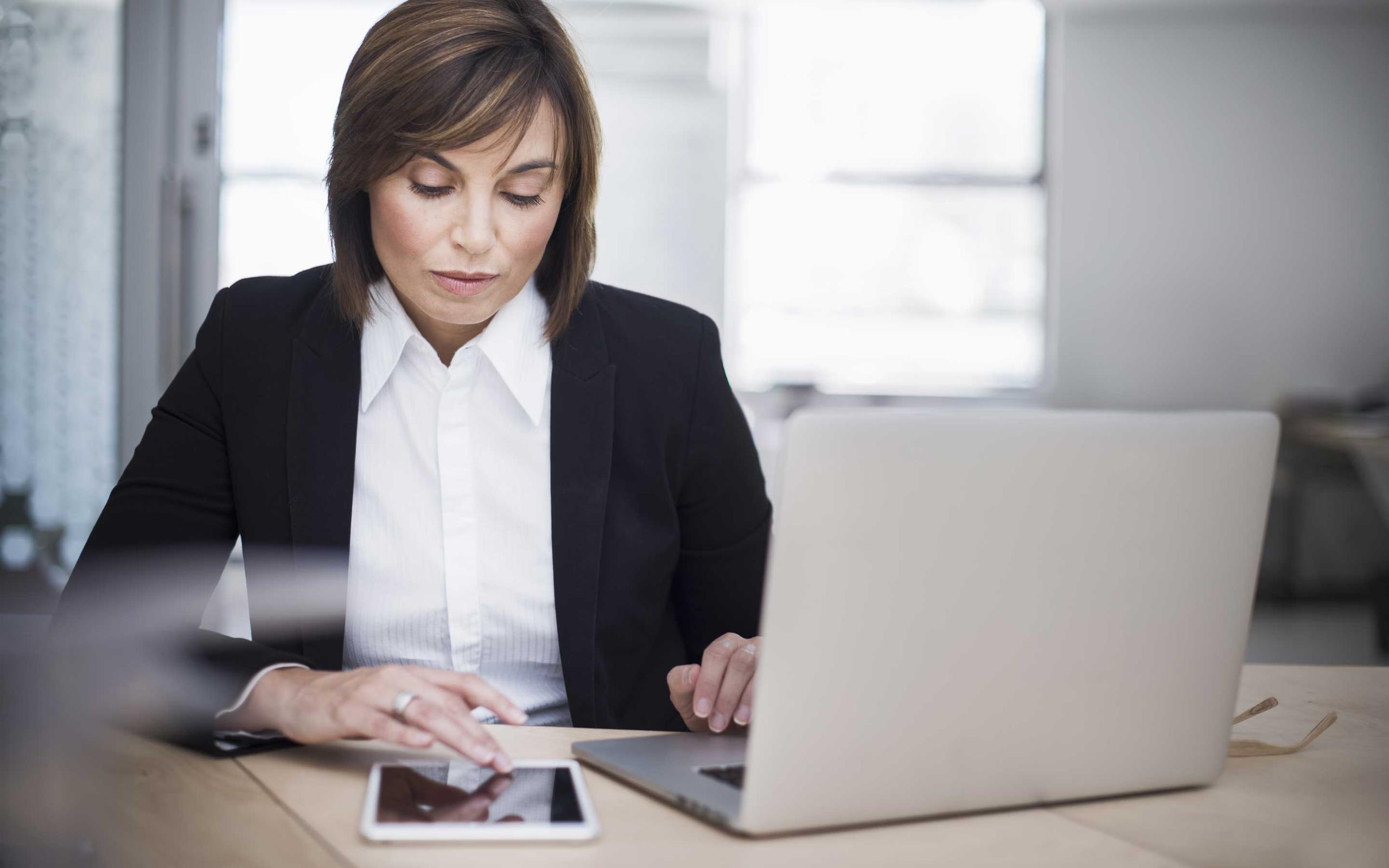 Woman sitting in front of laptop, looking at her tablet