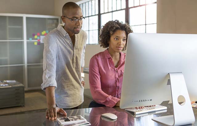Woman sitting at a desk looking at a large computer monitor while a man standing behind her looks at the monitor