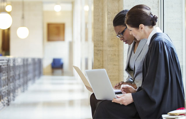 A judge and a lawyer sitting in a courtroom hallway looking at laptops