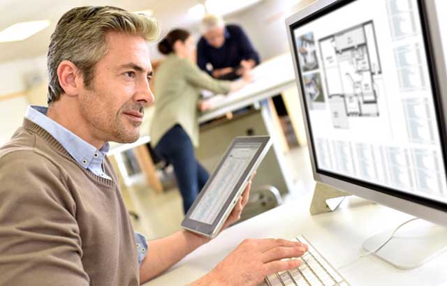 Man sitting at a desk working on a computer and holding a tablet while a man and woman are standing at a table in the background